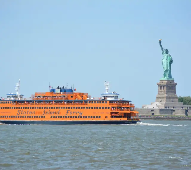 Pomarańczowy prom Staten Island Ferry płynie obok Statuy Wolności.