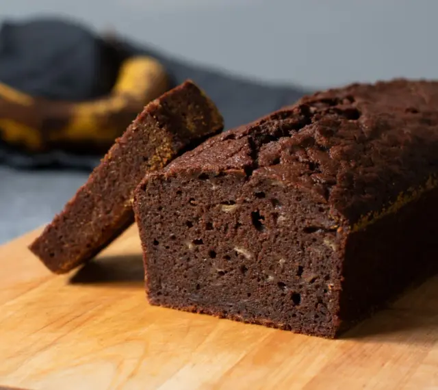 Chokladig banankaka, perfekt för ett enkelt banankaka recept. En skiva är uppskuren och en övermogen banan ligger i bakgrunden.