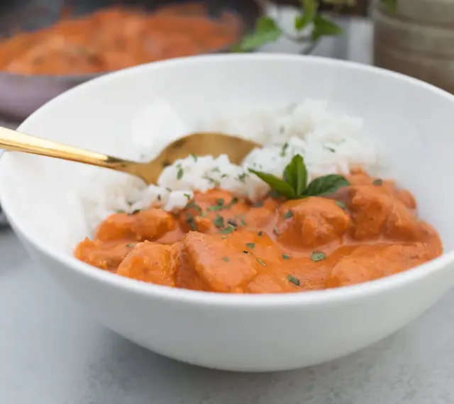 Aksamitny butter chicken masala z ryżem, udekorowany miętą. Złota łyżka zanurzona w daniu.