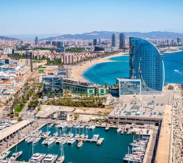 Barcelona morze: luksusowe jachty w marinie, plaża pełna ludzi i nowoczesny hotel w kształcie żagla.