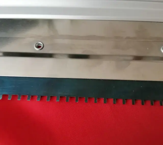 Metalowa rakla z ząbkowanym ostrzem, gotowa do pracy na czerwonym materiale.