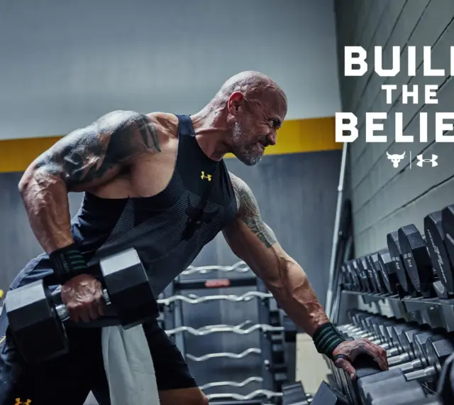 Dwayne "The Rock" Johnson w koszulce Under Armour wykonuje ćwiczenia z hantlami. W tle stojaki z ciężarami.
