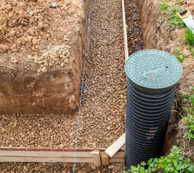 Budowa drenażu wokol domu. Wykop, żwir, rura drenarska i właz studzienki kanalizacyjnej.