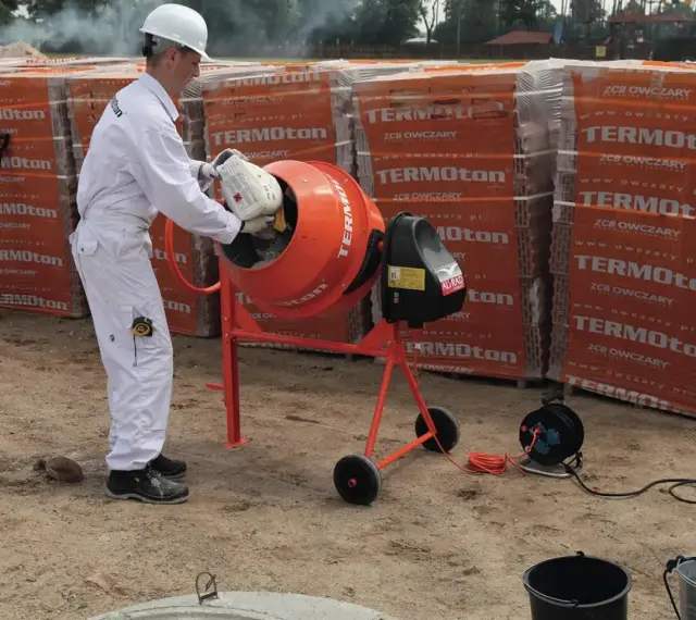 Budowlaniec w białym kombinezonie wsypuje składniki do betoniarki, przygotowując beton B20. W tle palety pustaków.