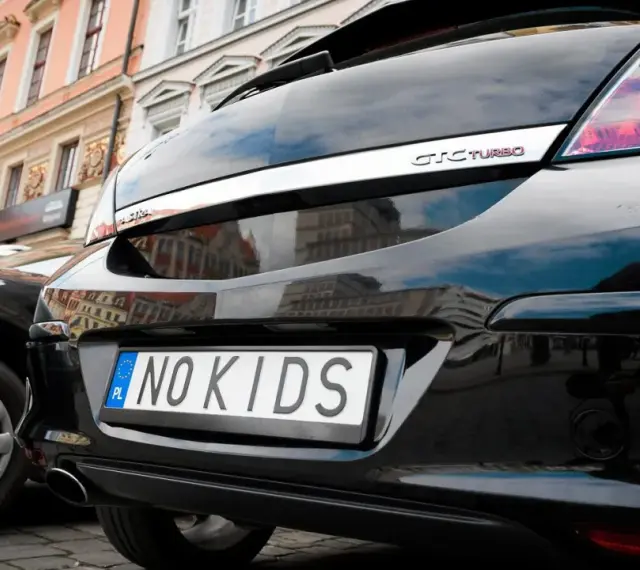 Czarne auto z indywidualnymi tablicami rejestracyjnymi "NO KIDS" na tle kamienicy.