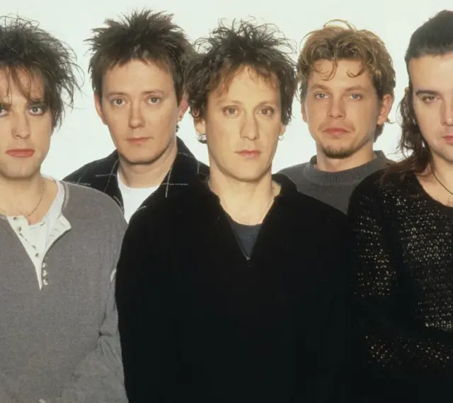 The Cure członkowie w szarych i czarnych ubraniach, z charakterystycznymi fryzurami i makijażem.