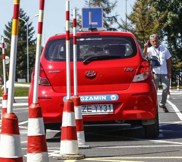 Czerwony Hyundai na placu manewrowym, z literą "L" na dachu. Instruktor pokazuje znak pokoju. Przygotowania do egzaminu na prawo jazdy b+e.