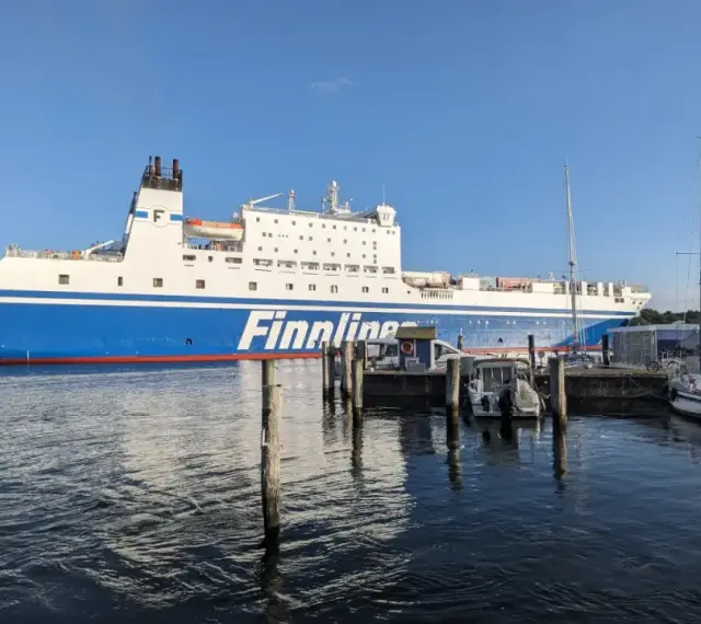 Duży prom Finnlines "Finntrader Malmö" cumuje w porcie, gotowy do rejsu. W tle widać żaglówki. Prom do Finlandii czeka na pasażerów.