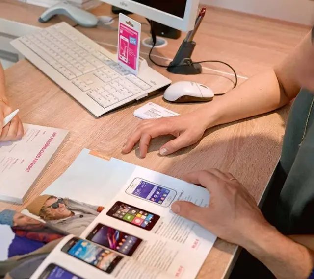 Dwie osoby przeglądają ofertę telefonów komórkowych. Na biurku leży klawiatura, myszka, skaner i katalog z produktami. Jedna z osób pisze coś na formularzu.