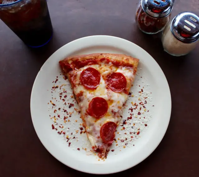 Kawałek pizzy pepperoni na talerzu, posypany płatkami chili i parmezanem. Idealny na szybki lunch, choć warto pamiętać o jego kcal.