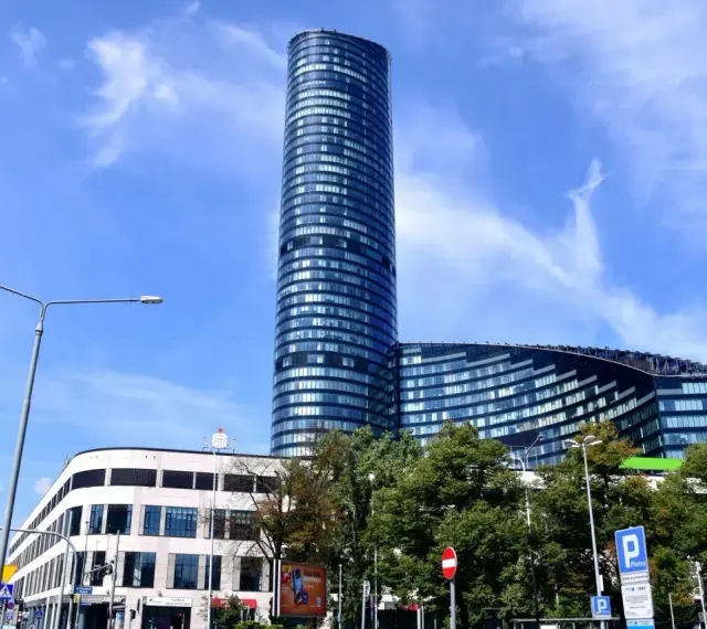 Wysoki, szklany budynek Sky Tower we Wrocławiu, z pewnością ma wiele pięter. Widać też niższy budynek i drzewa.