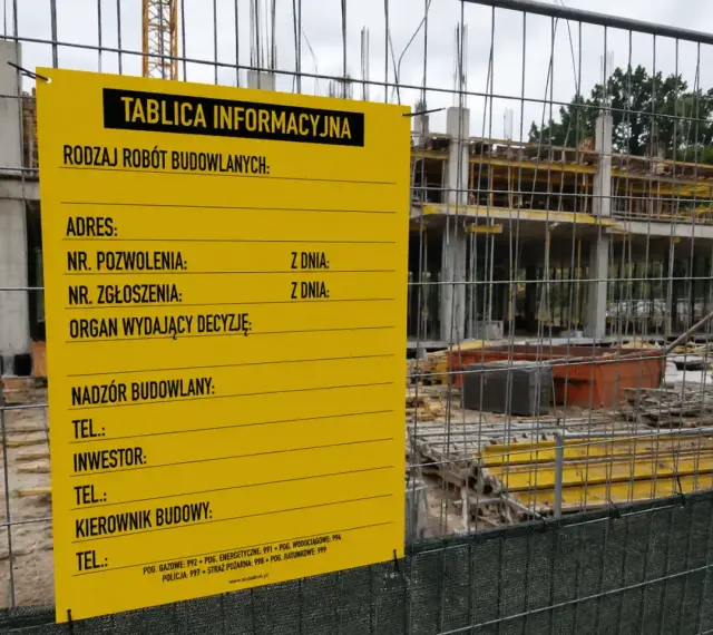 Żółta tablica informacyjna budowy z danymi o pozwoleniu, adresie i kontaktach. W tle widać konstrukcję budynku.