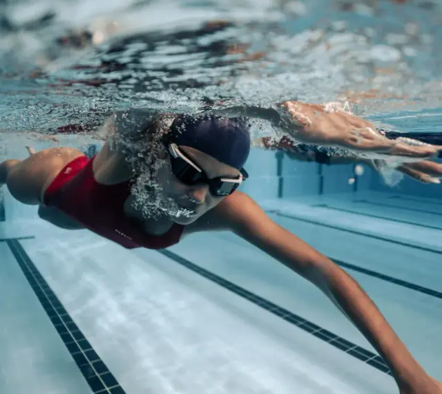Une femme en maillot de bain rouge nage en crawl, lunettes de natation sur le nez. Sa **natation transformation physique femme** est visible sous l'eau.