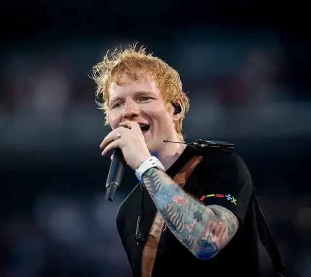 Ed Sheeran śpiewa z mikrofonem, jego ręka ozdobiona jest kolorowymi tatuażami.