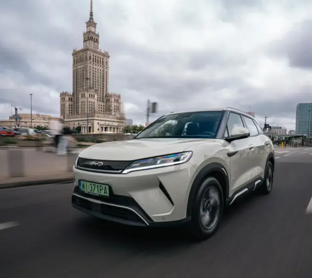 Biały, nowoczesny chiński samochód elektryczny BYD jedzie ulicą w Warszawie, z Pałacem Kultury w tle.