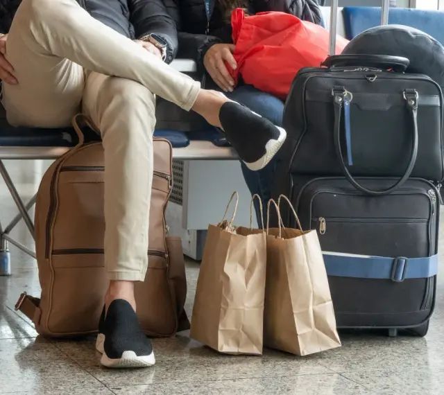 Osoby czekają na lotnisku z bagażem. Widać plecak, torby podróżne i papierowe torby na zakupy. To typowy nouvelair bagaż.