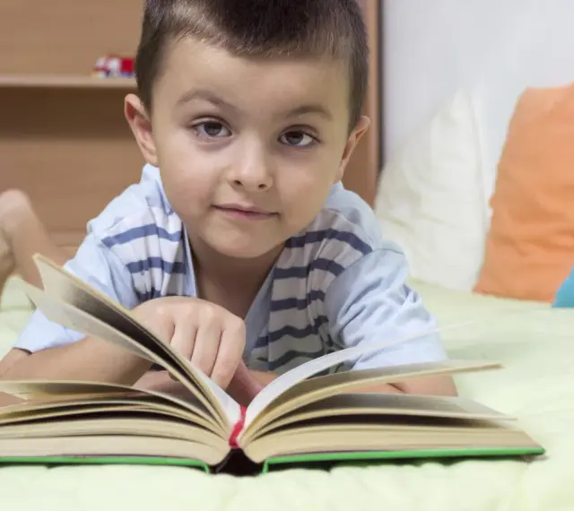 Chłopiec leży na łóżku z otwartą książką. To świetny sposób, jak zachęcić dziecko do czytania.