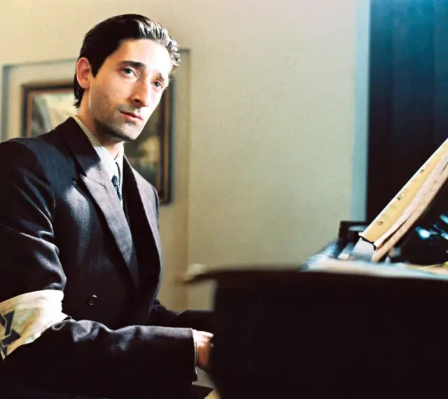 Adrien Brody jako Władysław Szpilman w filmie "Pianista" Romana Polańskiego, z opaską na ramieniu, siedzi przy fortepianie.