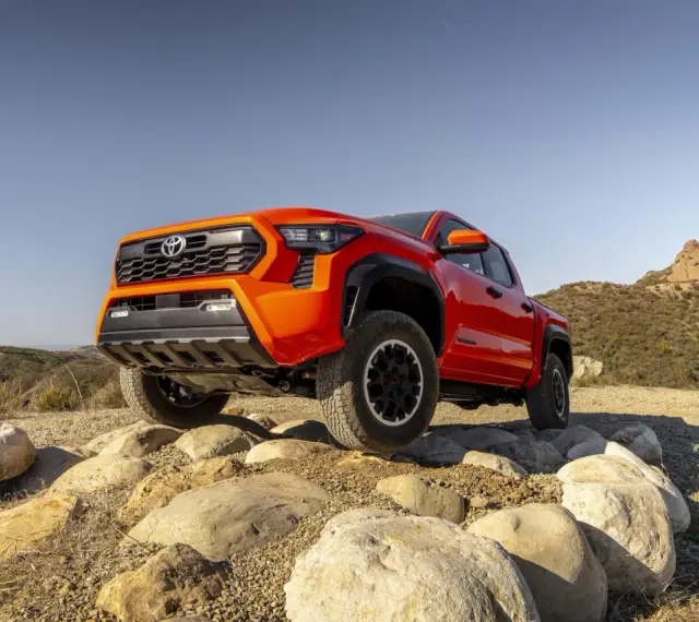 Pomarańczowy Toyota pickup pokonuje kamienisty teren na tle wzgórz.