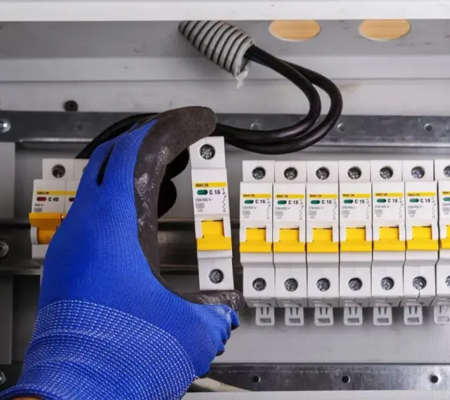 Ręka w niebieskiej rękawicy montuje bezpiecznik w skrzynce elektrycznej. Obok widać rząd innych bezpieczników i przewody. To ważny element instalacji, chroniący przed skutkami wyładowań, jak odgromnik.