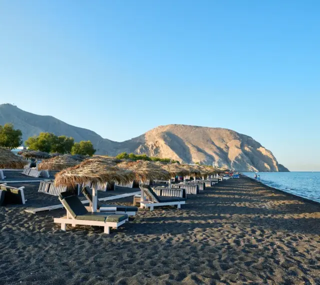 Czarne piaski i leżaki pod słomianymi parasolami na Santorini plażach. W tle góry i spokojne morze.