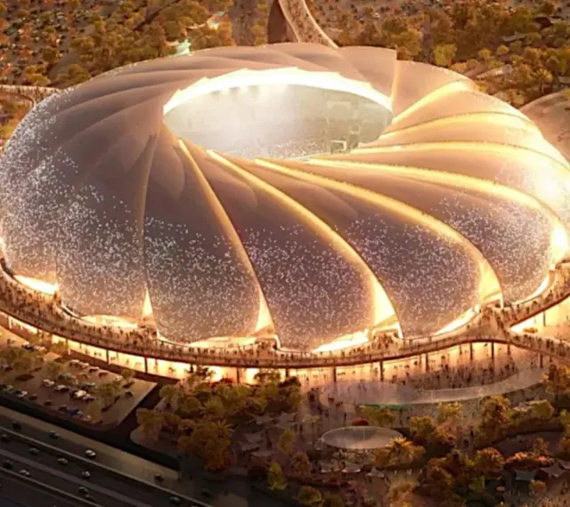 Futurystyczny stadion, który może być najładniejszy na świecie, z falującym dachem i oświetlonymi detalami, otoczony zielenią i tłumem ludzi.