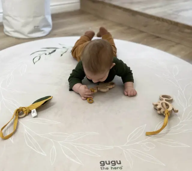 Niemowlę na brzuchu na macie z motywem roślinnym bawi się drewnianymi gryzakami.