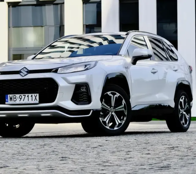 Biały Suzuki Across z czarnymi detalami stoi przed nowoczesnym budynkiem.