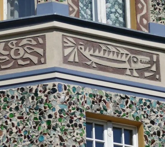 Sgraffito na fasadzie budynku: stylizowana ryba i abstrakcyjne wzory. Dolna część ozdobiona mozaiką z kolorowego szkła.