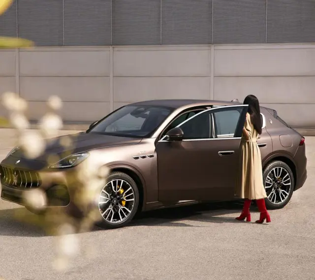 Elegancka kobieta w beżowej sukience i czerwonych kozakach zbliża się do brązowego Maserati SUV.