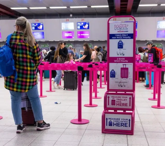 Kolejka do odprawy Wizzair. Tablica informuje, co trzeba wyjąć z bagażu podręcznego: płyny, elektronikę i przedmioty ostre.