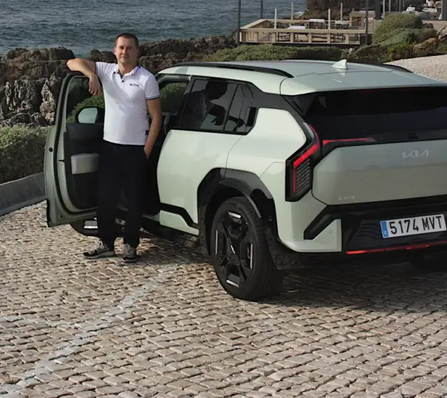 Mężczyzna opiera się o drzwi nowego, jasnoszarego Kia EV3, zaparkowanego na brukowanej drodze nad morzem.