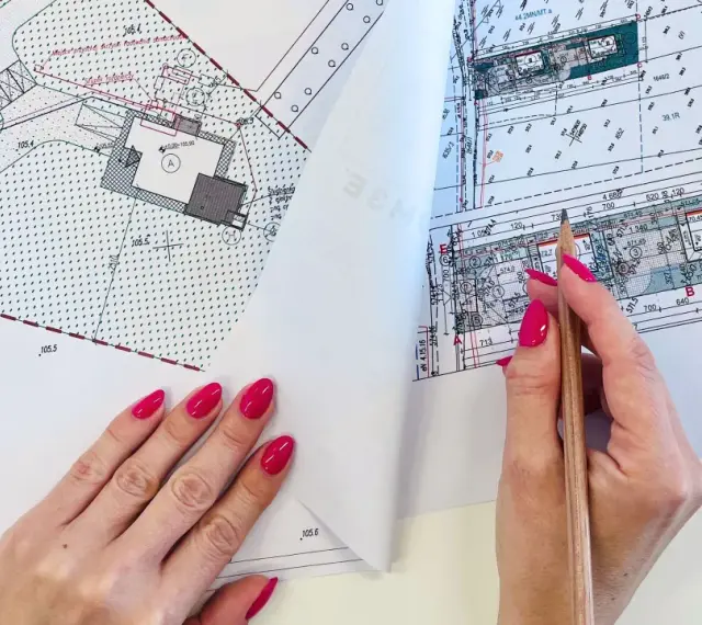 Ręka z różowymi paznokciami wskazuje ołówek na mapie do celów projektowych, gdzie widoczne są plany budowy i przyłączy.