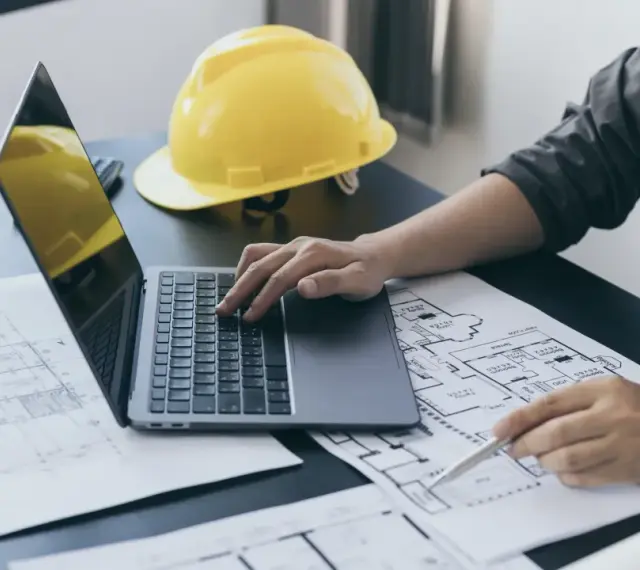 Architekt pracuje nad projektem, analizując plany i obliczając powierzchnię całkowitą budynku. Żółty kask ochronny leży obok.
