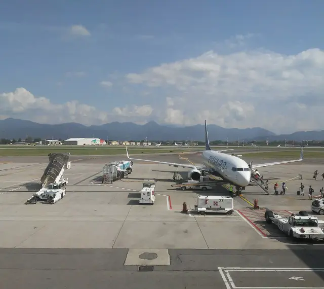 Samolot Ryanair na płycie lotniska Bergamo. W tle góry, na pierwszym planie schody, pojazdy obsługi naziemnej i pasażerowie.
