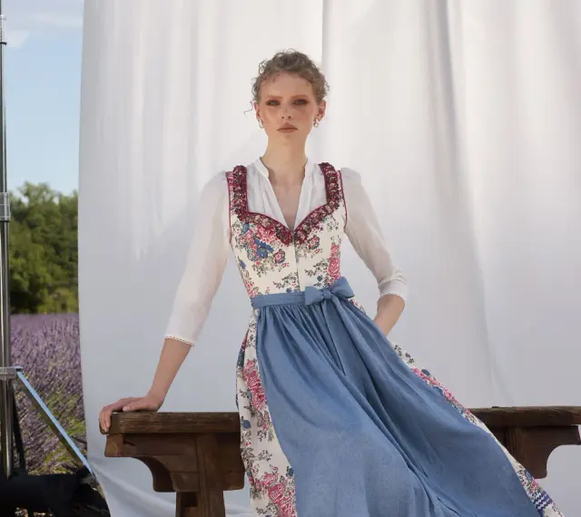 Ein Dirndl im floralen Muster mit blauem Rock, ein moderner Dirndl-Trend, posiert vor Lavendelfeld-Kulisse.