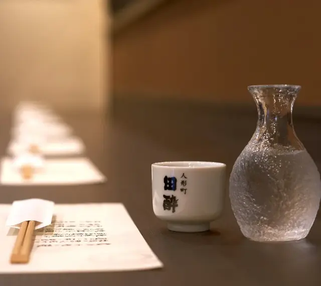 Sake ile procent? Delikatna czarka i karafka z sake czekają na degustację. W tle rząd nakryć.