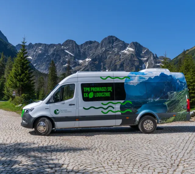 Biały bus Tatrzańskiego Parku Narodowego z grafiką górskiego krajobrazu, przypominającego Morskie Oko, stoi na kamiennej drodze.