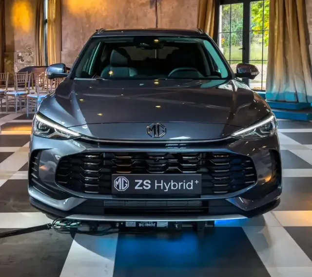 Szary MG ZS Hybrid+ prezentuje się elegancko na tle wnętrza z zasłonami i krzesłami.