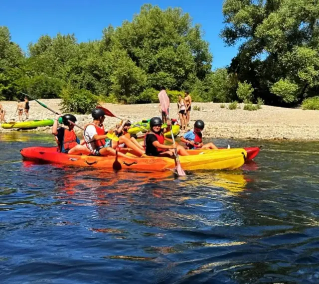 Des personnes profitent d'une activité canoe kayak sur une rivière ensoleillée, entourées de verdure et d'une plage de galets.