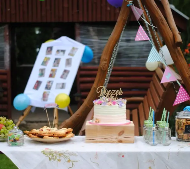 Na stole na roczek kładzie się tort, owoce, ciasteczka i słodkości. Dekoracje tworzą girlandy i zdjęcia.