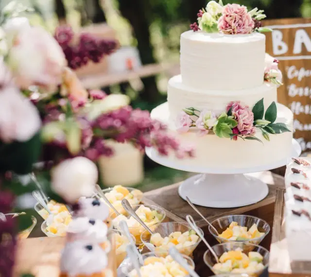 Elegancki tort weselny boho ozdobiony kwiatami, obok deserki i owoce.