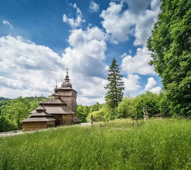 Drewniana cerkiew na tle błękitnego nieba z chmurami, otoczona zielenią. Widok z łąki w Beskidzie Dolnym.