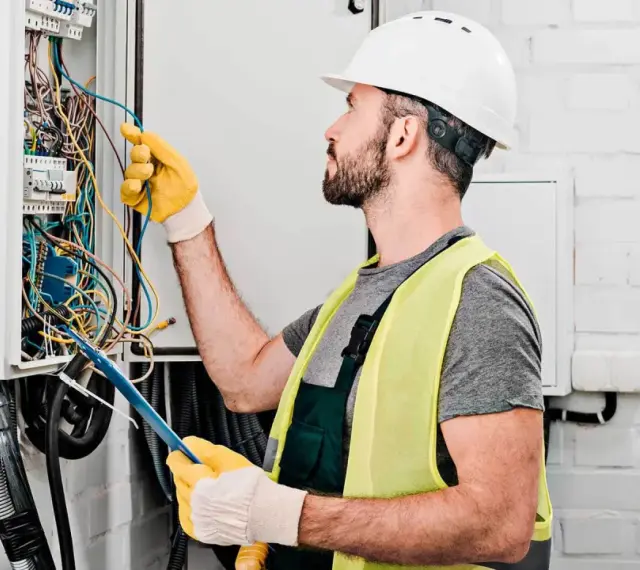 Elektryk w kasku i kamizelce odblaskowej pracuje przy skrzynce z przewodami. Sprawdź nasz cennik usług elektrycznych!