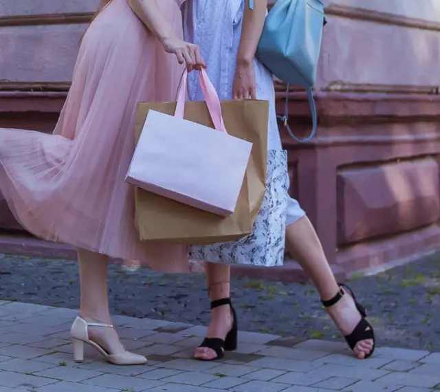 Dwie kobiety w zwiewnych sukienkach i modnych, szerokich obcasach w damskich butach, niosą torby z zakupami.