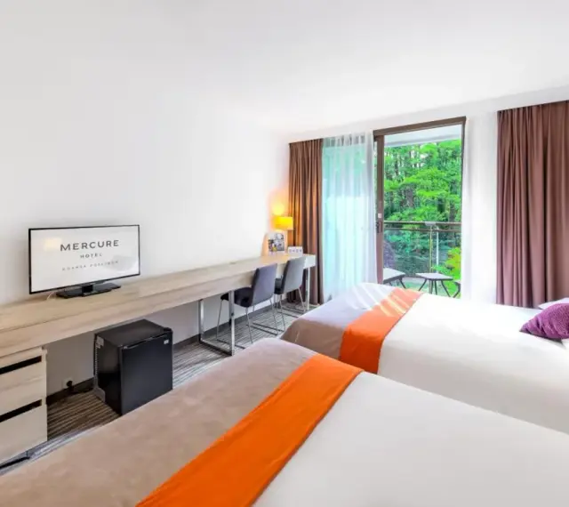 Pokój hotelowy z dwoma łóżkami, biurkiem i balkonem z widokiem na zieleń. Hotel Posejdon Gdańsk.