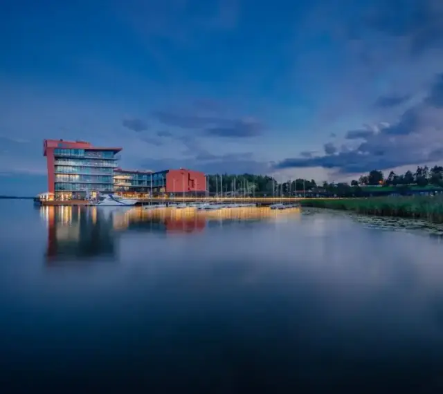 Nowoczesny hotel na ptasiej wyspie, otoczony spokojną wodą i zielenią, z łodziami przy pomoście.