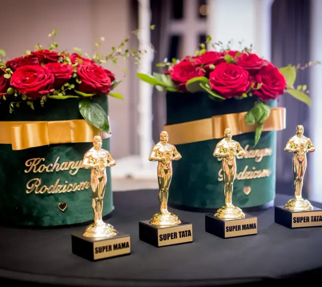 Złote statuetki "Super Mama" i "Super Tata" obok bukietów róż. Różnica między podziękowaniami dla rodziców w formie filmu a na żywo? Te są na żywo!