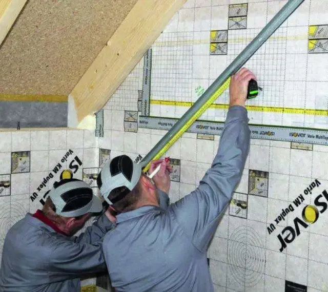 Dwóch budowlańców pracuje przy konstrukcji szkieletowej. Jeden mierzy taśmą, drugi zaznacza.