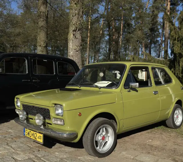Zielony fiat 127 z lat 70. stoi obok czarnej taksówki na tle drzew.